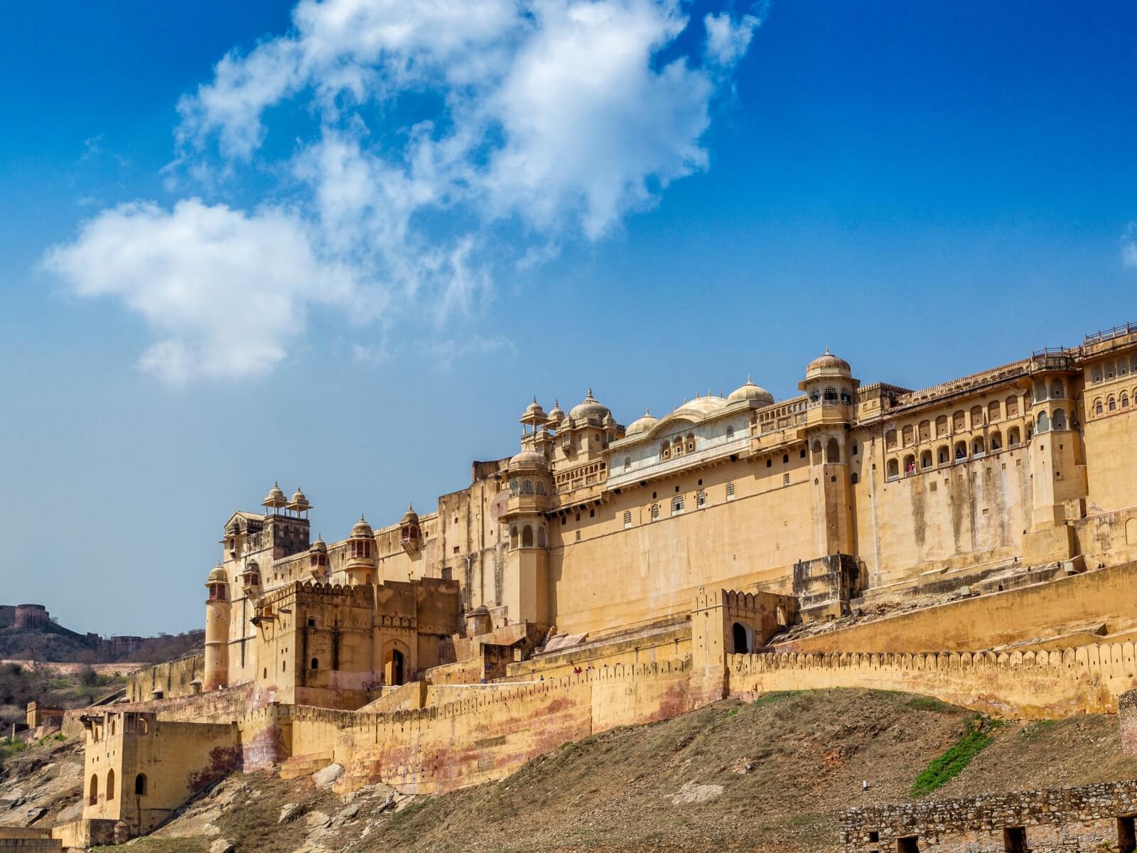 A majestic exterior view of the imposing yellow sandstone walls, intricate domes, and sprawling architecture of Amber Fort Jaipur, rising prominently from a rugged hillside under a bright blue, cloud-dotted sky.