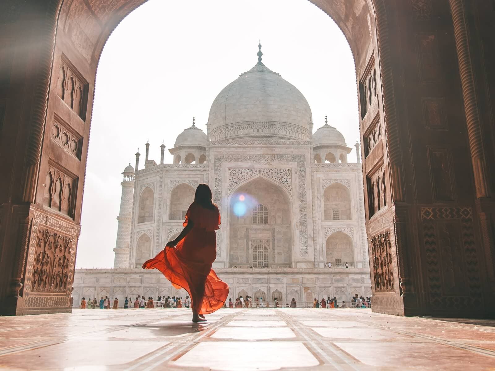A woman in a flowing orange dress stands framed by a carved stone archway, looking out at the iconic Taj Mahal—a majestic highlight often included in North India tour packages.