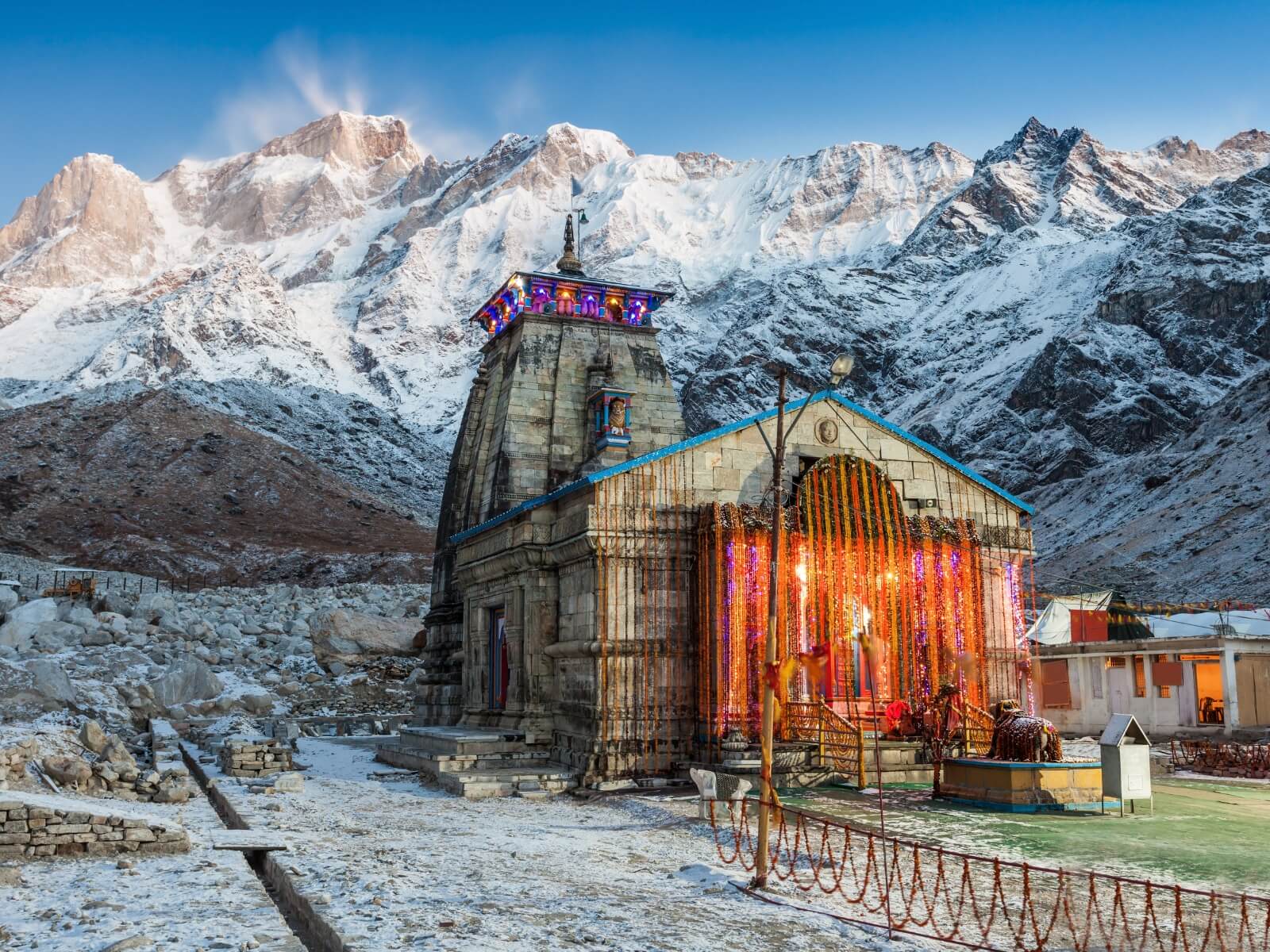 The beautifully illuminated Kedarnath Temple, adorned with bright lights and vibrant orange garlands, stands majestically amidst a snowy landscape against a striking backdrop of towering, snow-capped Himalayan peaks—a deeply spiritual and breathtaking destination often highlighted in Uttarakhand Tour Packages.