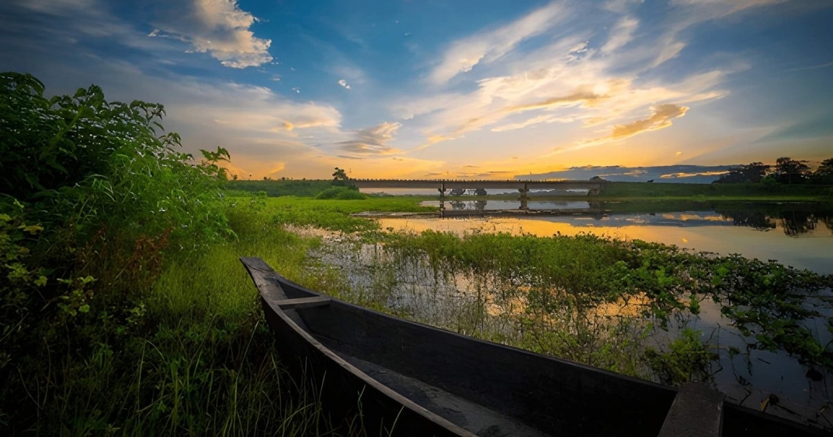 A rustic wooden boat moored in the lush green wetlands of Majuli Island, Assam, overlooking a calm river that perfectly reflects a vibrant, cloudy sunset and a distant bridge.