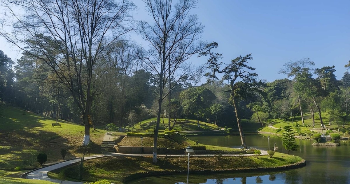 A peaceful park setting featuring a calm lake surrounded by manicured grassy mounds, meandering paved walking paths, and tall trees, some with bare branches indicating the autumn season.