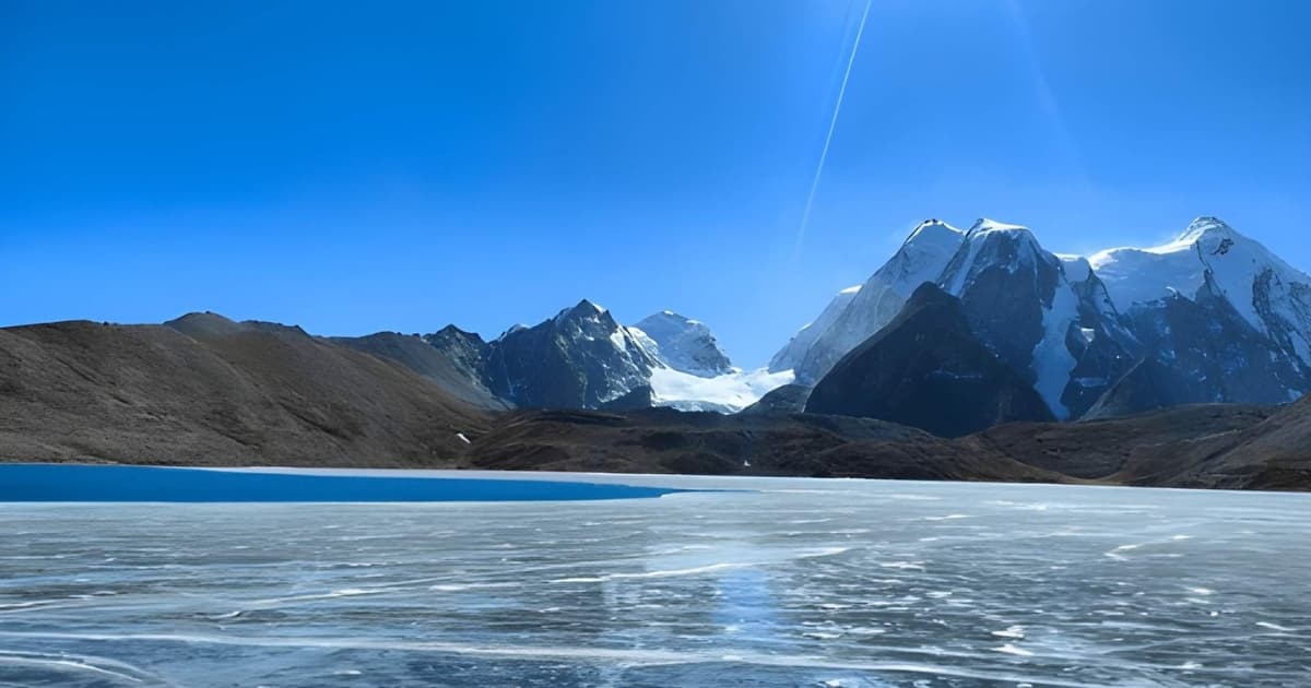 A wide view of a high-altitude, partially frozen lake. Intricate white ice formations cover the foreground, transitioning into a patch of deep blue water, with majestic, sharply defined snow-covered mountains towering under a bright blue sky.