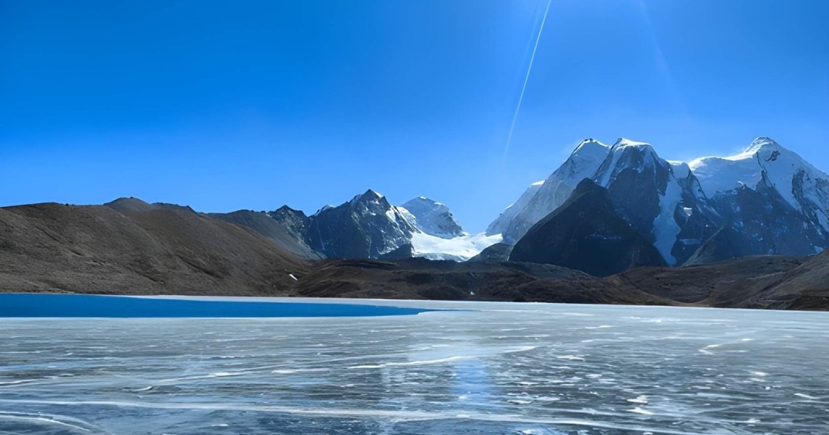 A wide view of a high-altitude, partially frozen lake. Intricate white ice formations cover the foreground, transitioning into a patch of deep blue water, with majestic, sharply defined snow-covered mountains towering under a bright blue sky.
